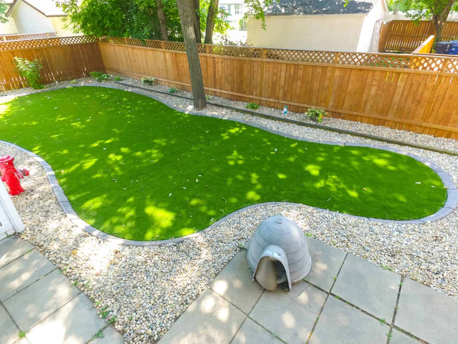 Backyard with kidney-shaped artificial grass, surrounded by decorative stones, wooden fence, and a small dog house.