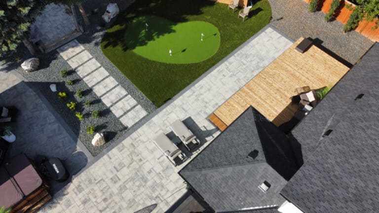 Aerial view of a modern backyard featuring a light wood deck, a grey stone paver patio, a kidney-shaped synthetic turf putting green, and a hot tub.